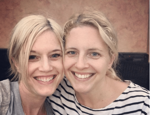 Two women with blonde hair smiling, sitting closely together for a selfie in front of a brown background.