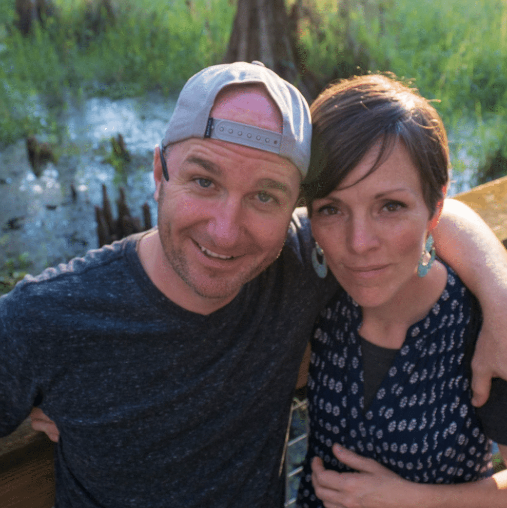 A man in a backwards cap and a woman in a patterned shirt stand close together outdoors, with greenery and a pond in the background.