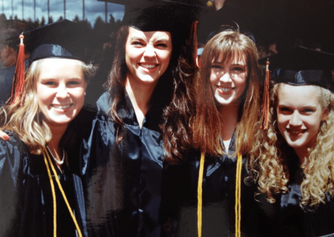 Four young women in graduation caps and gowns stand close together, smiling outdoors at a graduation ceremony.