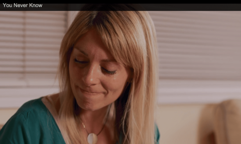 A woman with blonde hair looks down and smiles slightly while sitting indoors. The text "You Never Know" appears at the top of the image.