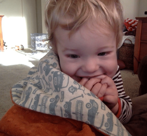 A young child with blonde hair holds a blue-and-white patterned blanket and smiles while sitting indoors on a carpeted floor.