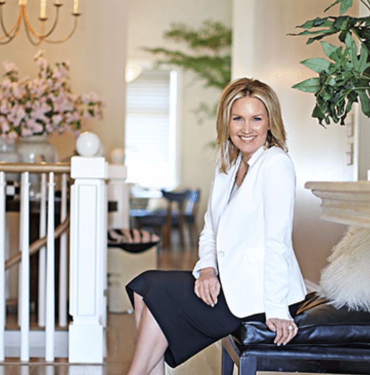A woman in a white blazer and black skirt sits on a bench in a well-decorated, modern living space with plants and flowers in the background.