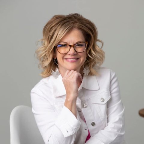 Woman with short, wavy blonde hair and glasses wearing a white jacket sits on a chair, smiling with her hand resting under her chin against a plain background.