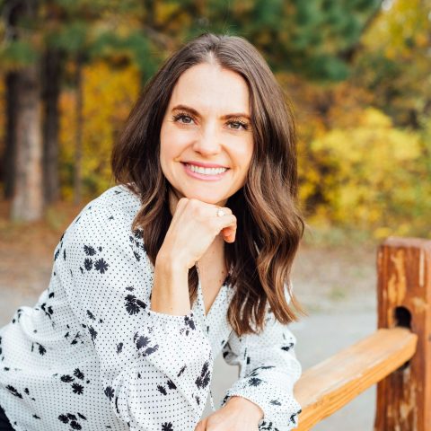 A woman with long brown hair wearing a white shirt with black patterns smiles while leaning on a wooden fence outdoors with trees in the background.