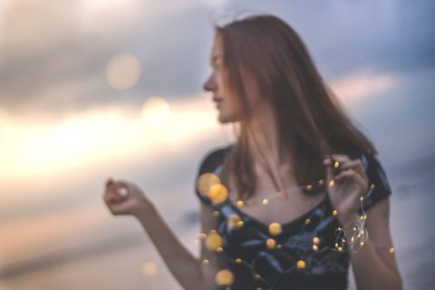 A woman stands outdoors holding string lights, with a blurred background and soft focus on her and the lights.