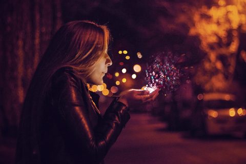 A person in a leather jacket holds their hands near their face, blowing colorful particles into the air on a dimly lit street at night.