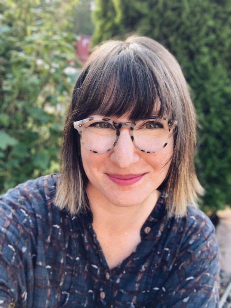 A person with shoulder-length brown hair, glasses, and a patterned shirt smiles outdoors in front of greenery.
