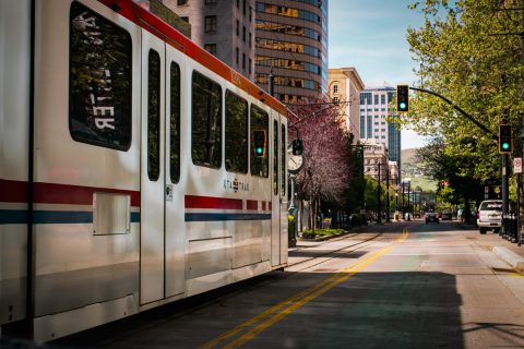 A white light rail train travels through a city street lined with trees and tall buildings on a clear day, with traffic lights showing green.