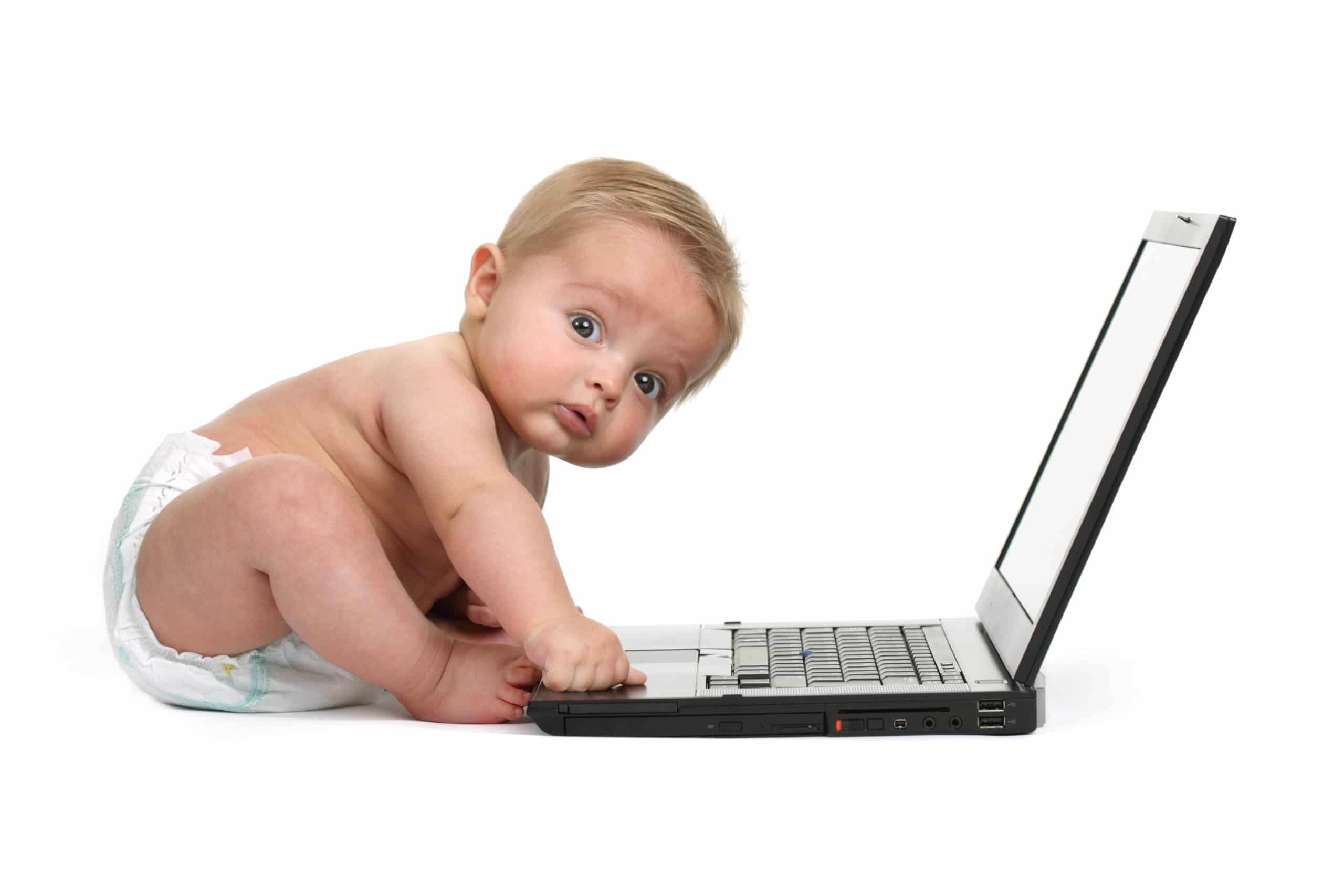 A baby in a diaper sits on the floor and presses a key on a laptop, looking up with a curious expression. The background is plain white.