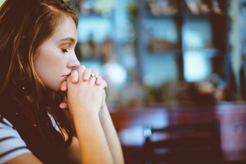 A woman sits indoors with her eyes closed and hands clasped together, appearing deep in thought or prayer.