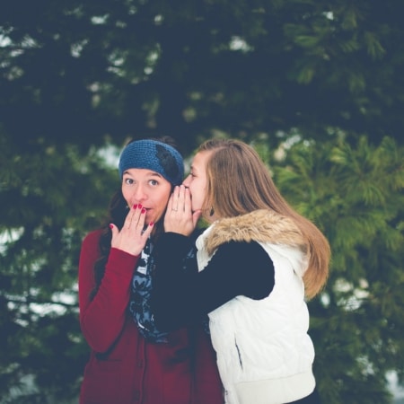 Two women standing outside; one whispers into the other’s ear while the other looks surprised, covering her mouth with her hand. Evergreen trees are visible in the background.