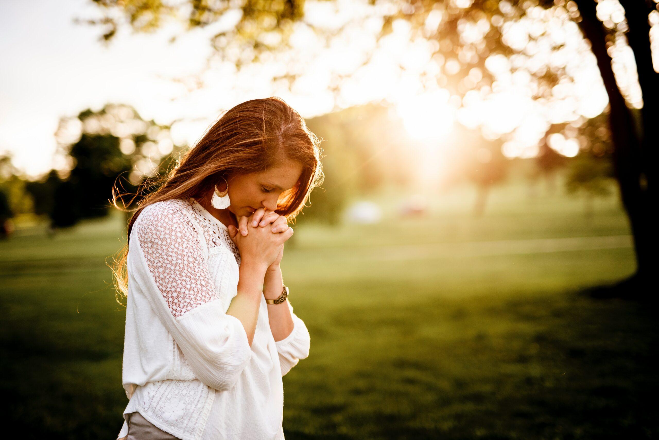 A woman stands outdoors in a sunlit park, with her head bowed and hands clasped together in front of her face, possibly in prayer or contemplation.