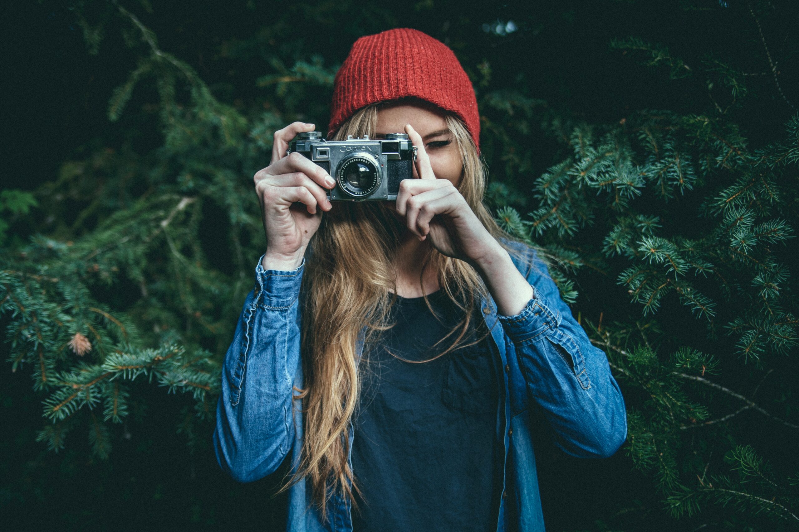 A person wearing a red beanie and denim jacket holds a vintage camera and takes a photo in front of green foliage.