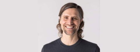 Thomas McConkie, with long brown hair, a trimmed beard, and a grey t-shirt, smiles at the camera against a plain light background.