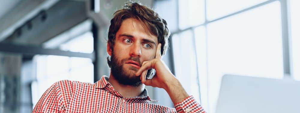 A man with a beard, wearing a red checkered shirt, holds a phone to his ear and looks passive-aggressive and concerned while sitting indoors near a window.