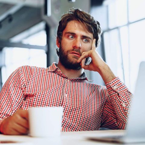 Bearded man in a red checkered shirt sits at a desk, holding a white mug and looking at his laptop screen with a thoughtful, almost passive-aggressive expression.