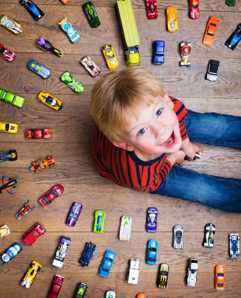 A young boy sits on a wooden floor, smiling up at the camera, surrounded by an organized circle of colorful toy cars.