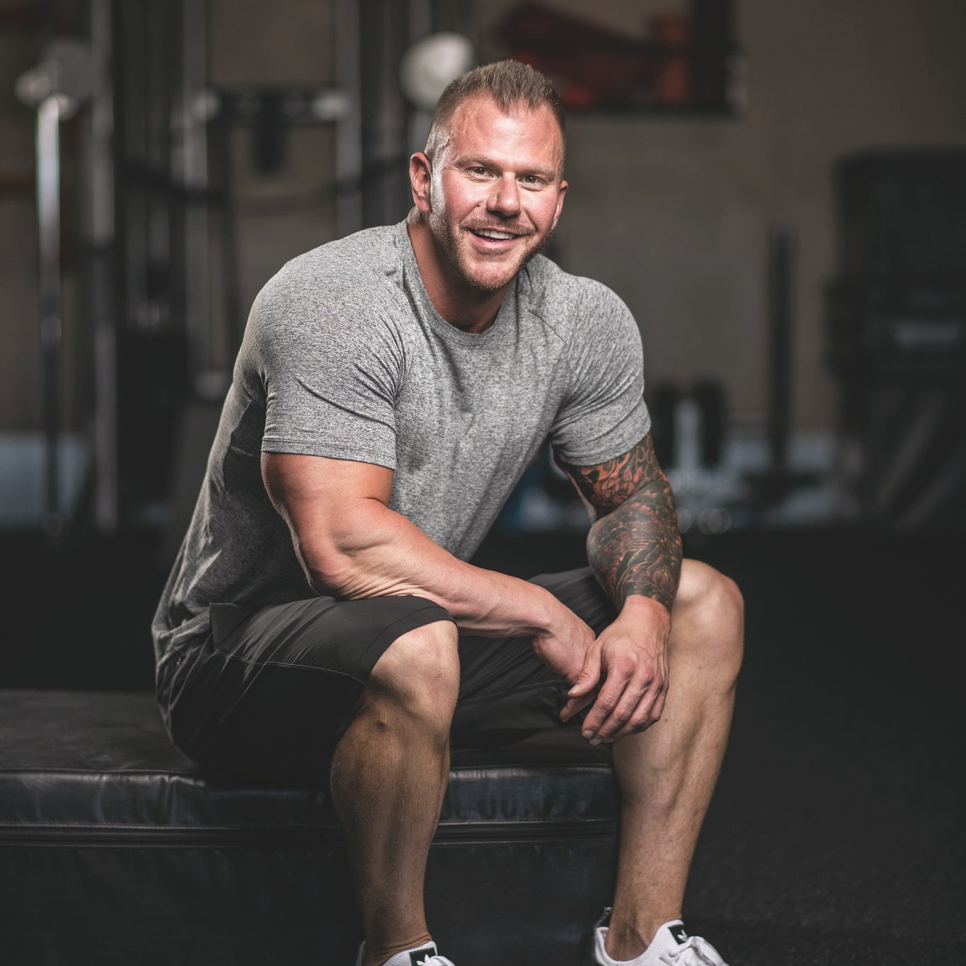 A man with short hair, wearing a grey t-shirt and black shorts, sits on a gym bench, smiling at the camera. Gym equipment is visible in the background.