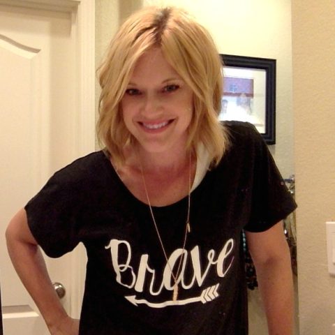 Woman with blonde hair smiles at the camera, wearing a black t-shirt with the word "Brave" and an arrow graphic, standing indoors near a door and framed picture.