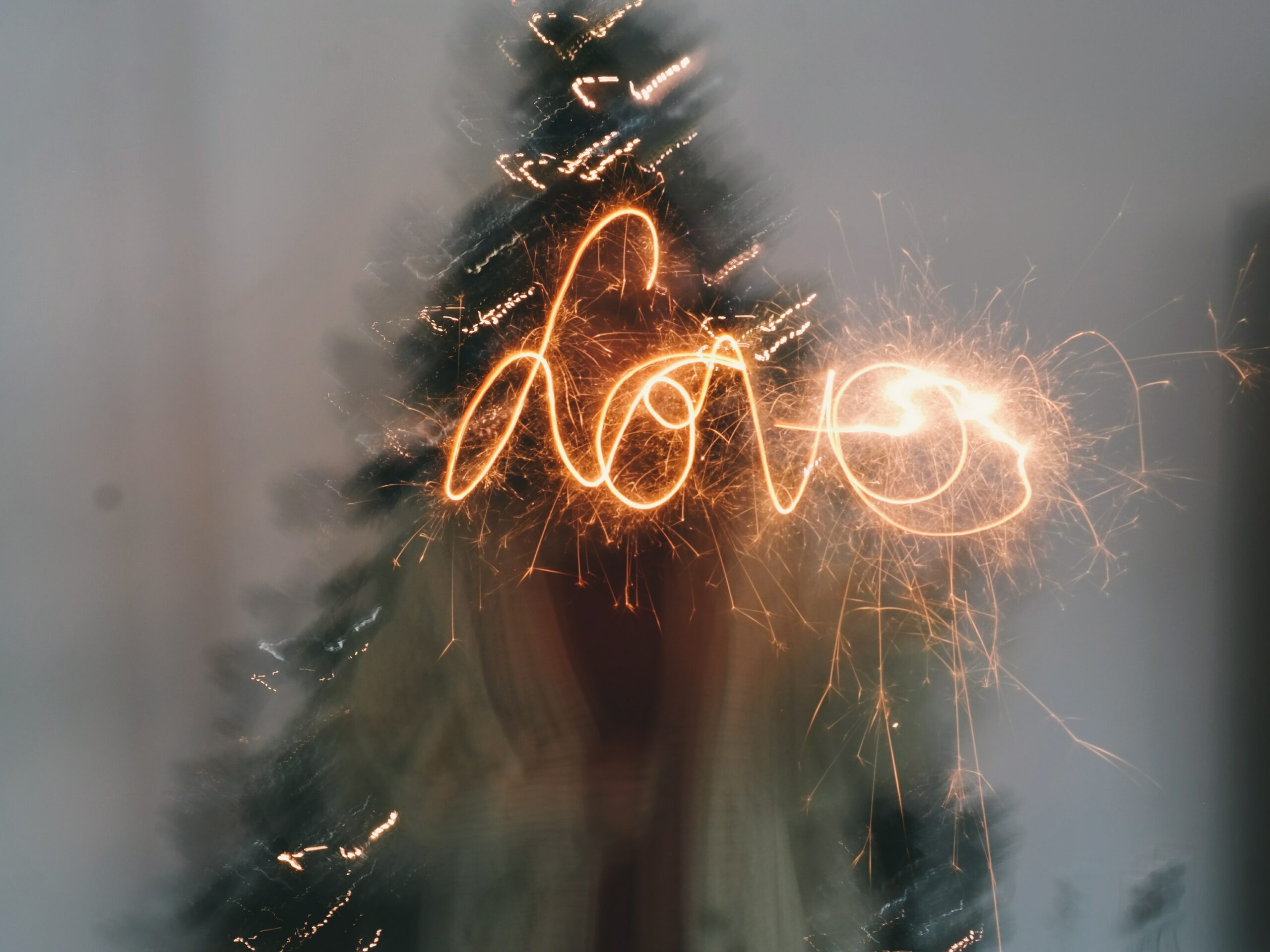 A person stands in front of a Christmas tree, using a sparkler to write the word "love" in the air, creating a bright light trail effect.