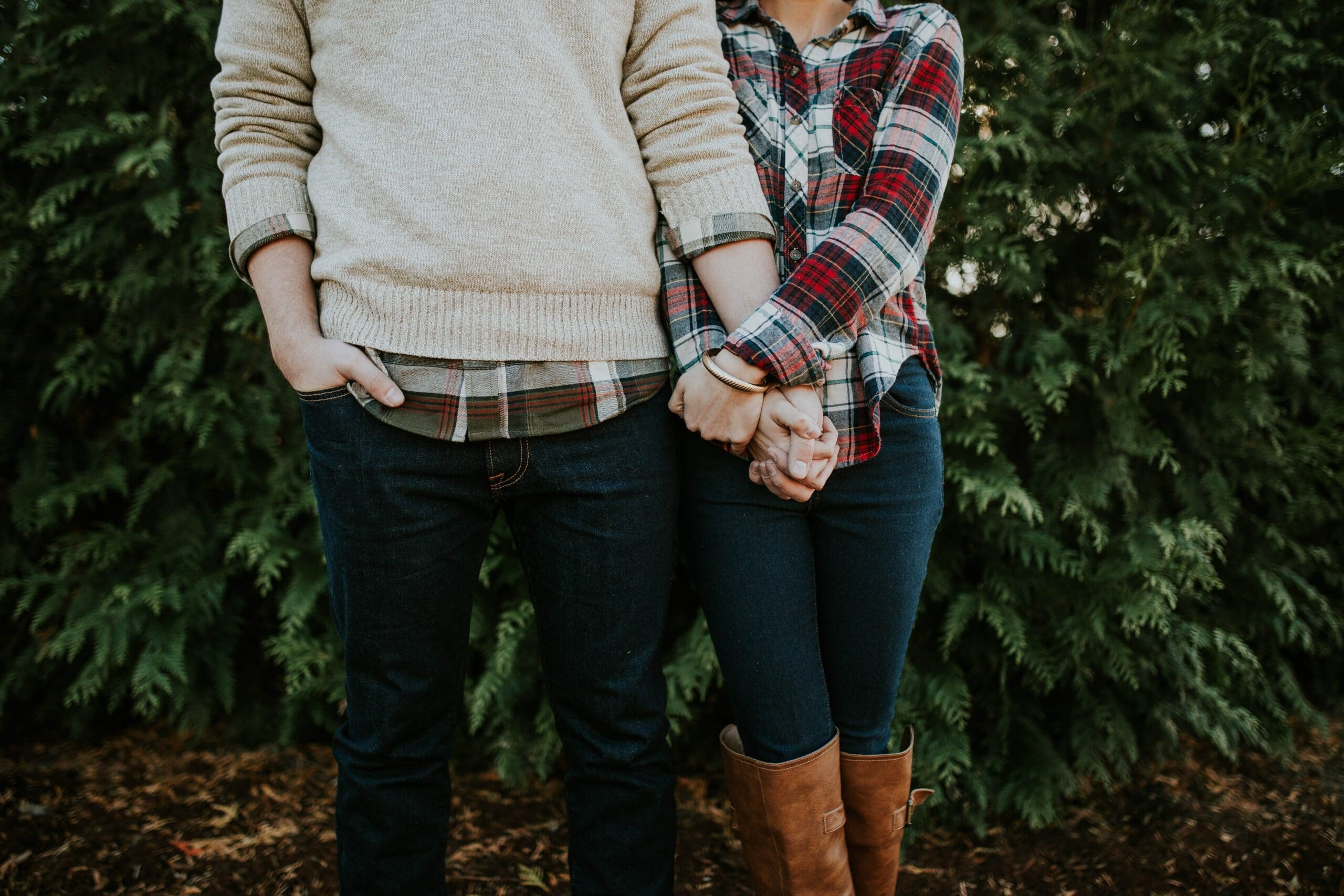 Two people standing side by side outdoors, holding hands. Both are wearing jeans; one has a plaid shirt and boots, the other a beige sweater. Green foliage in the background.
