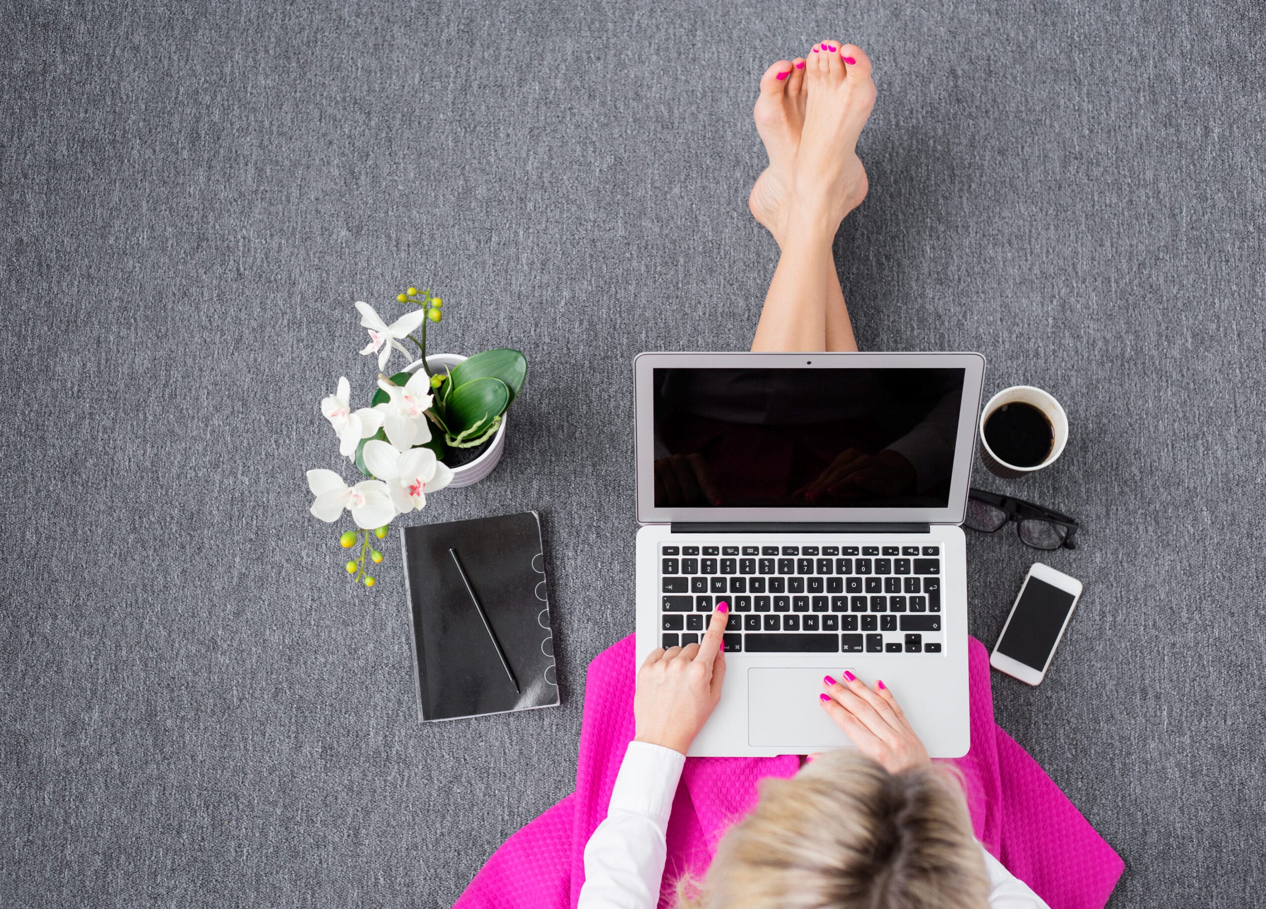 Top view of a person using a laptop on a gray carpet, with a notebook, pen, coffee cup, smartphone, and a potted orchid nearby.