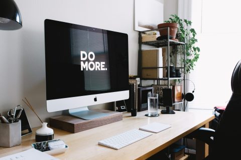 A desktop computer on a wooden desk displays the text "DO MORE." The desk holds office supplies, a glass of water, and potted plants, with shelves and a window in the background.