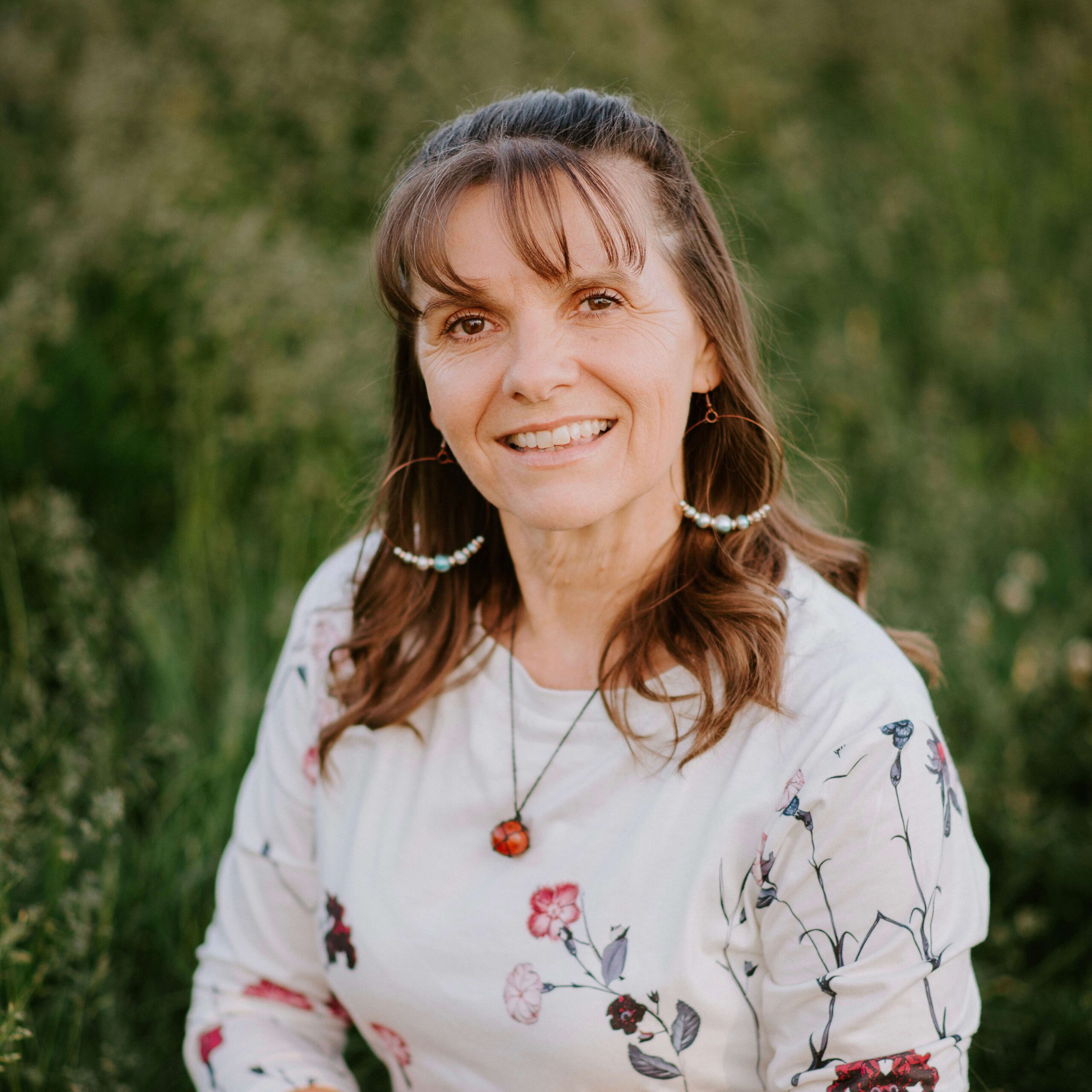 Woman with long brown hair and hoop earrings, wearing a floral white shirt, sits outdoors in a field of tall green grass, smiling at the camera.