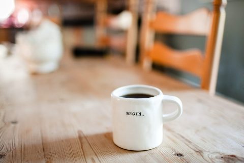 A white coffee mug with the word “BEGIN.” printed on it sits on a wooden table with chairs in the background.