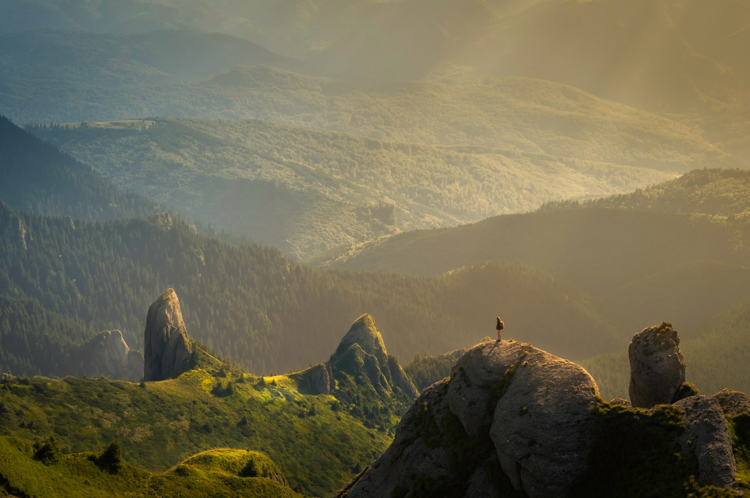 A person stands on a rocky peak overlooking rolling green hills and forested mountains under soft sunlight.