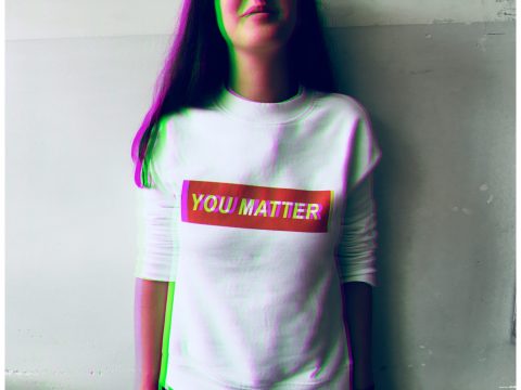 A person stands against a plain wall wearing a white long-sleeve shirt with "YOU MATTER" printed in bold letters on the front.