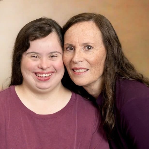 Two women with long brown hair, one younger and one older, both wearing purple tops, pose closely together and smile at the camera against a neutral background.