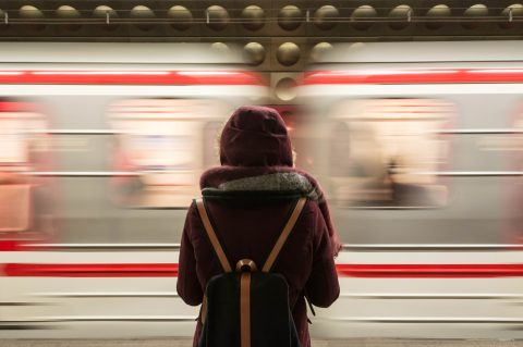 A person wearing a hooded coat and backpack stands on a subway platform as a train speeds by in the background.