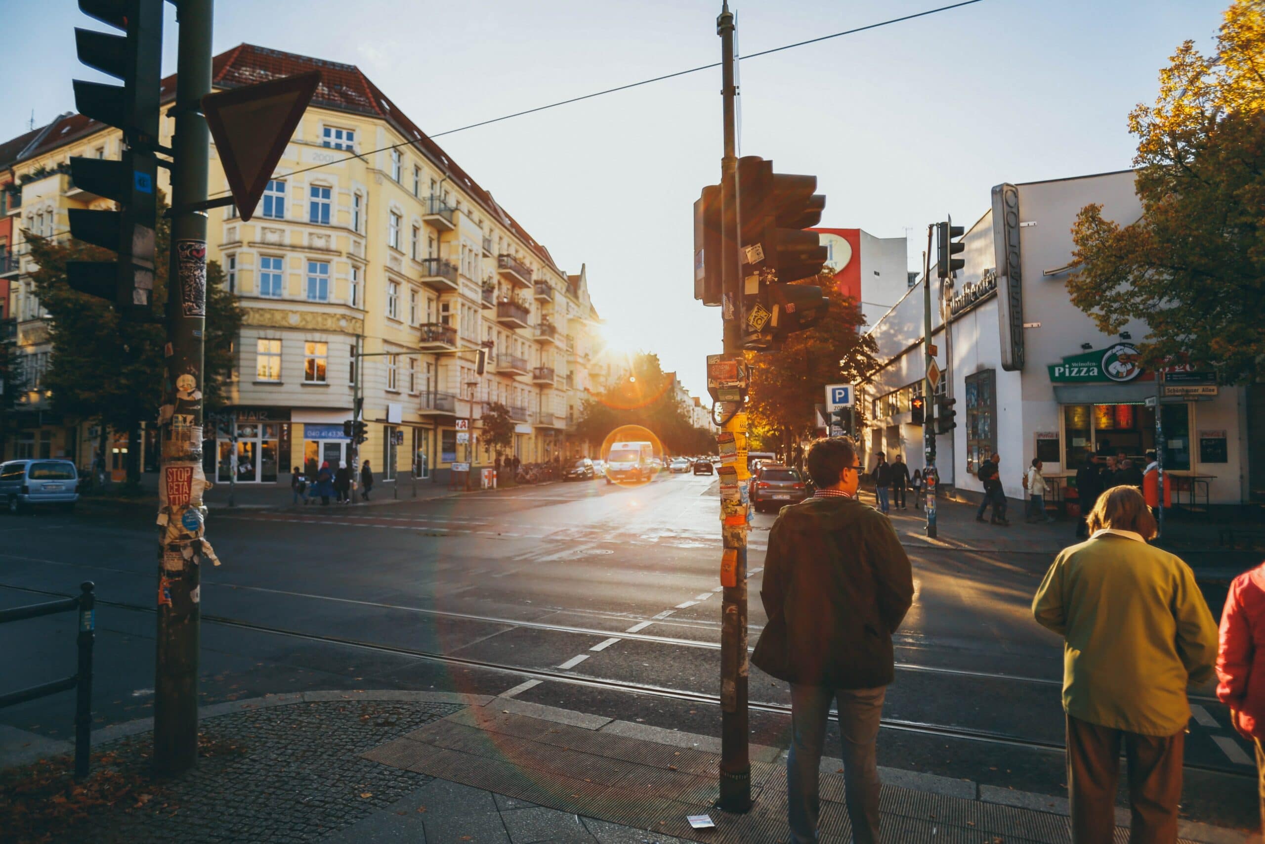 People wait at a crosswalk on a city street corner as the sun sets, with cars and buildings in the background.