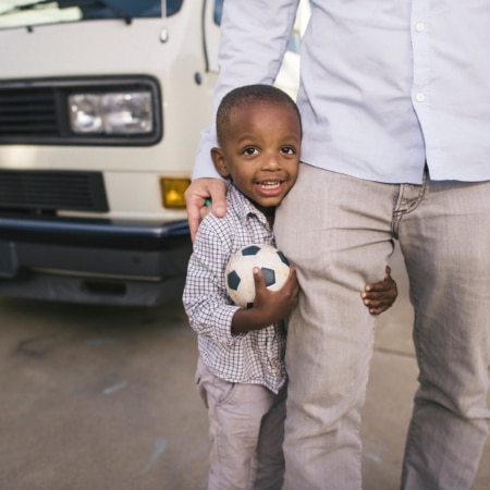 A young boy holding a small soccer ball stands next to an adult, partially hidden, in front of a white van.