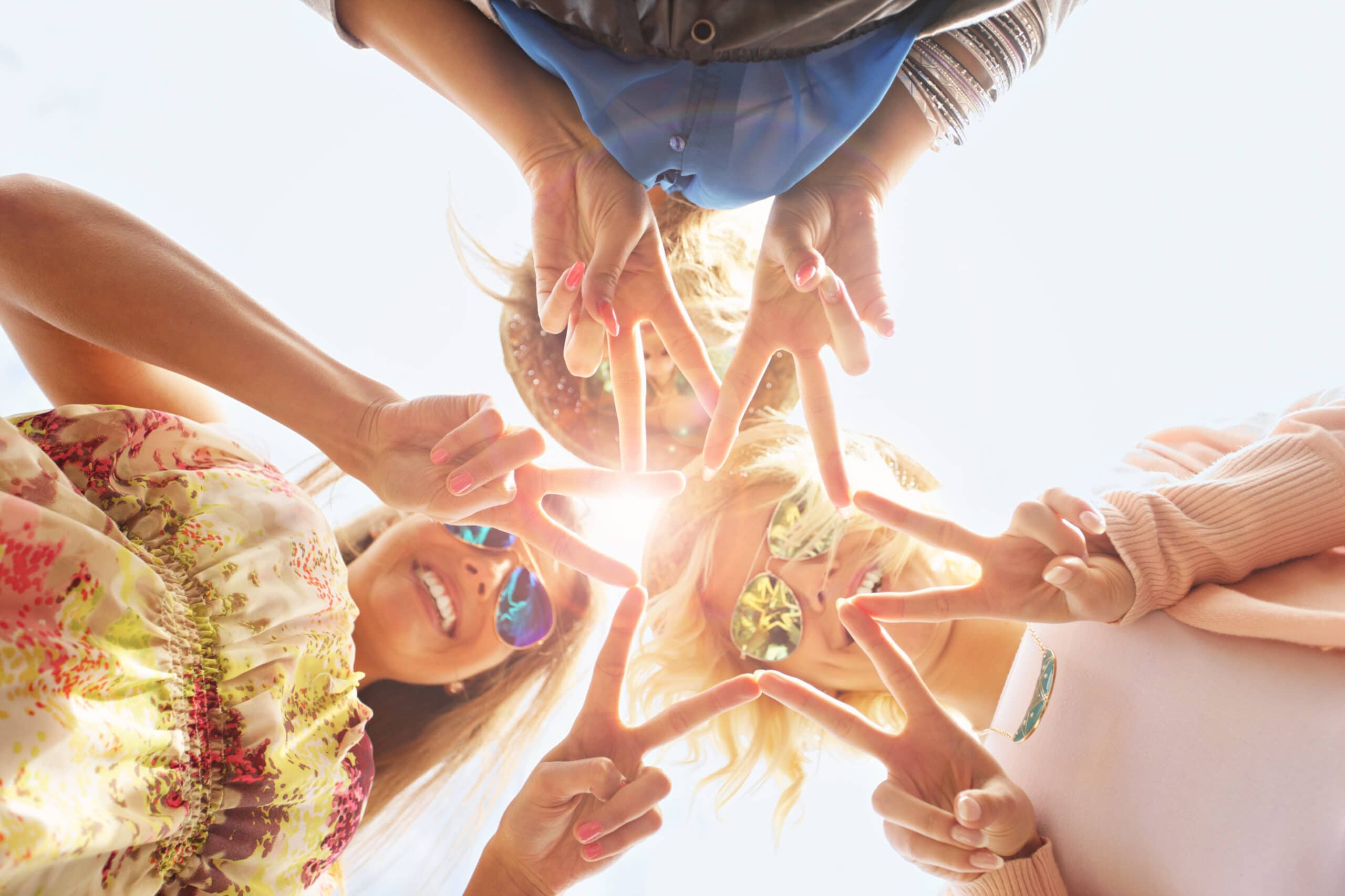 Three people standing in a circle outdoors, looking down at the camera, making a star shape with their fingers. Sunlight shines through their hands.