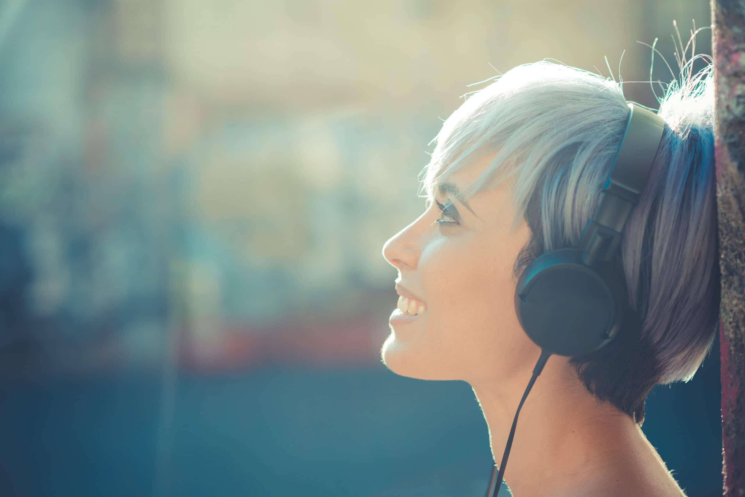 Person with short, light hair wearing black headphones, smiling and looking upward, with a blurred outdoor background.