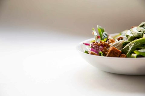 A white bowl filled with a fresh salad containing greens, red onions, lentils, and croutons sits on a light surface with a plain background.