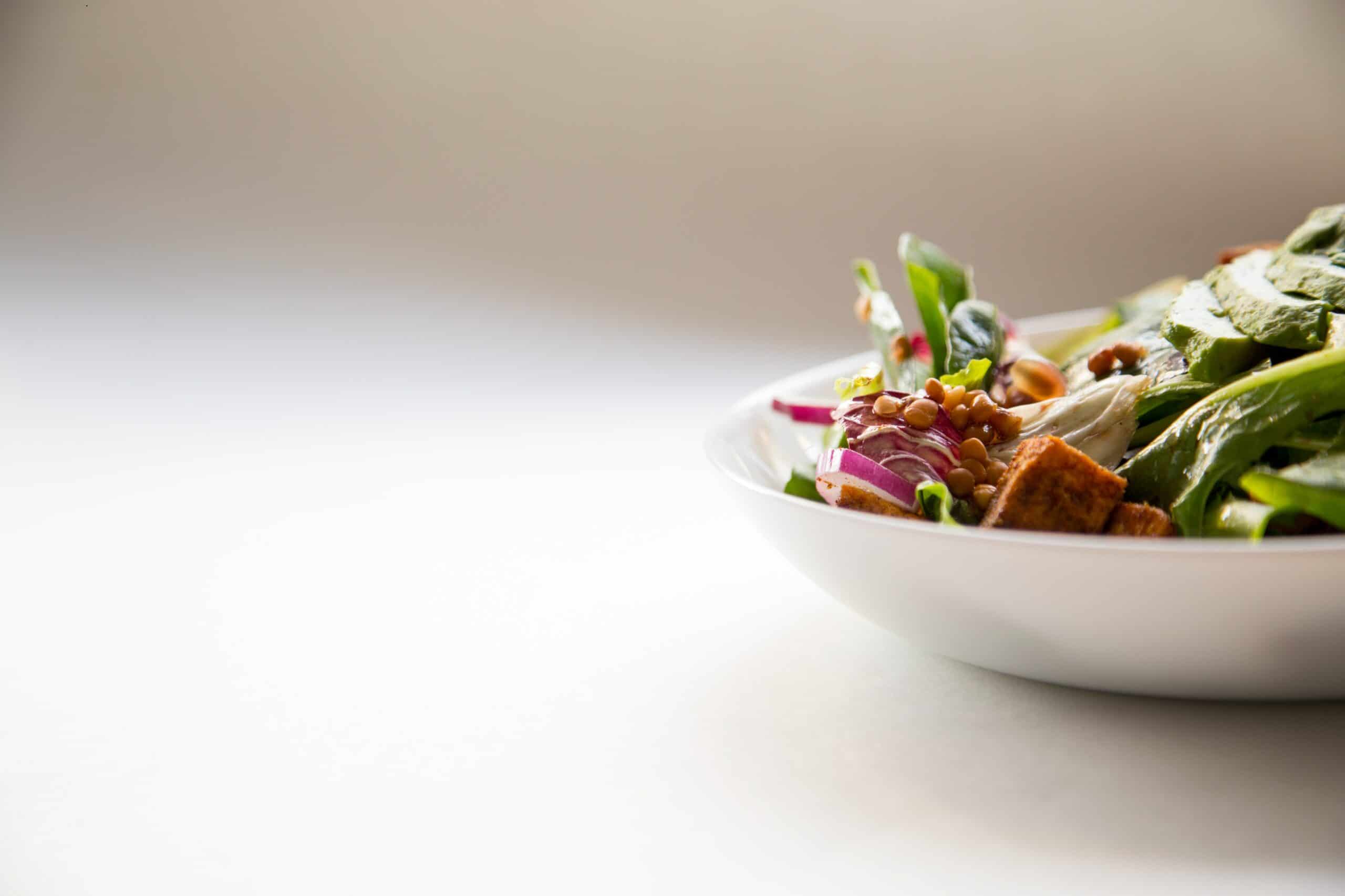 A white bowl filled with a fresh salad containing greens, red onions, lentils, and croutons sits on a light surface with a plain background.