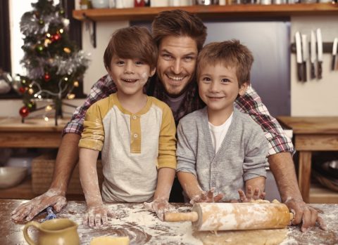 An adult and two children smile at the camera in a kitchen, with flour on their hands and a rolling pin on the counter, and a decorated Christmas tree in the background.