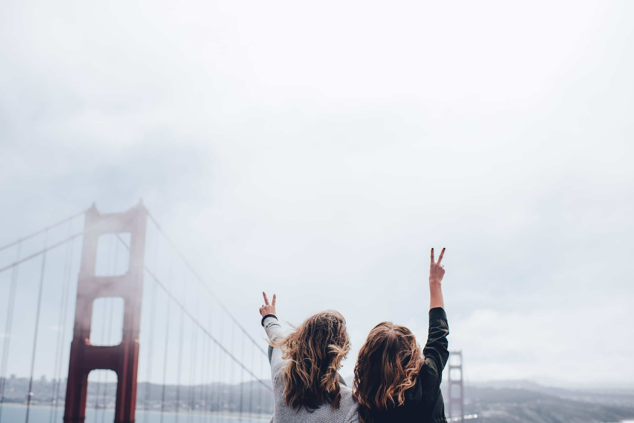 Two people stand with their backs to the camera, raising peace signs, in front of the Golden Gate Bridge on a cloudy day.