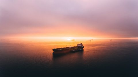 Cargo ships on calm water during sunrise, with the sky and sea reflecting orange and purple hues.