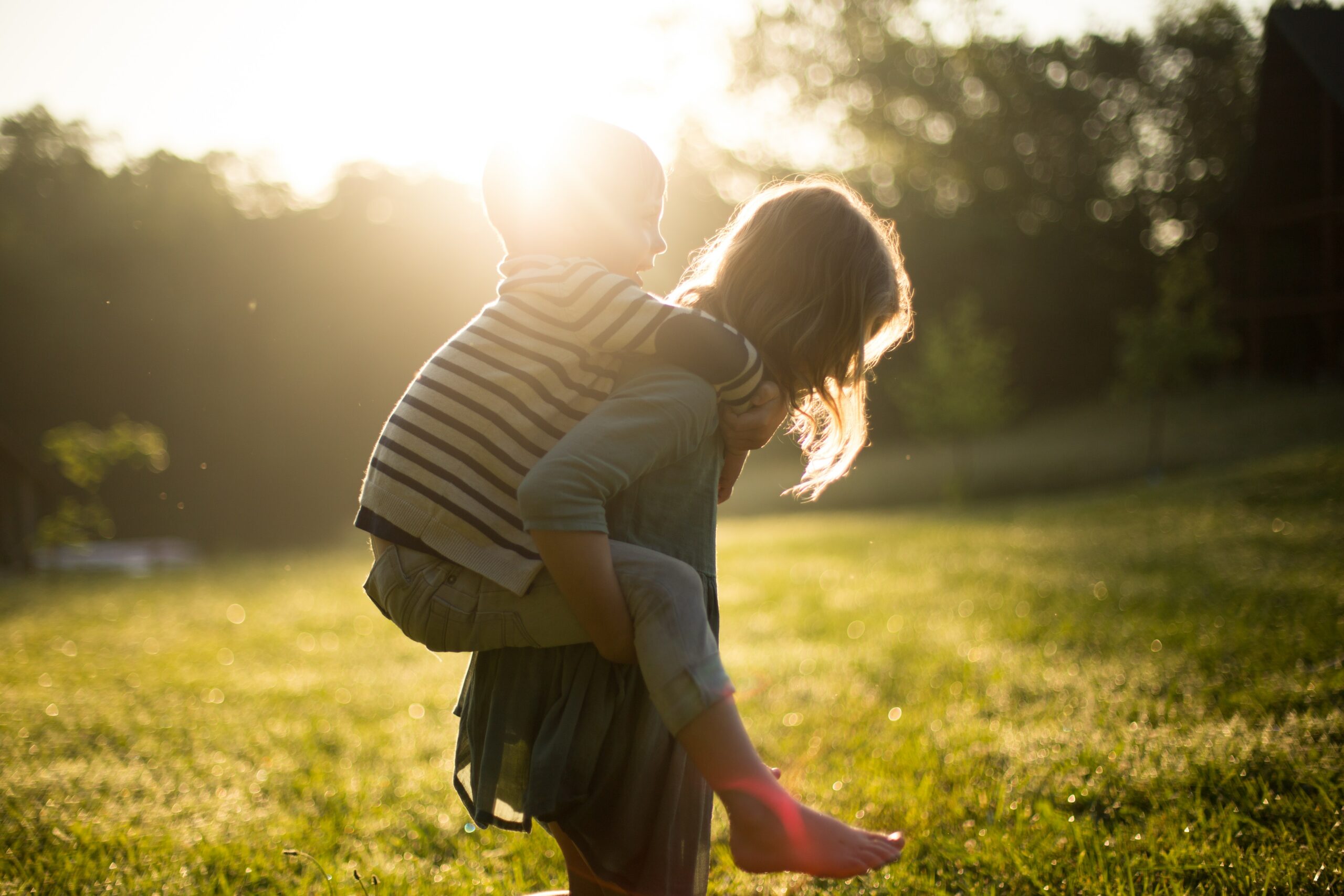 A child gives another child a piggyback ride outdoors on a grassy field with sunlight in the background.
