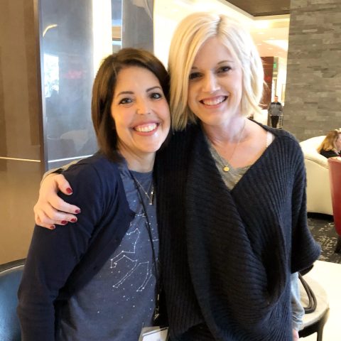 Two women are standing side by side indoors, smiling at the camera. One woman has her arm around the other. They both have short hair and are casually dressed.