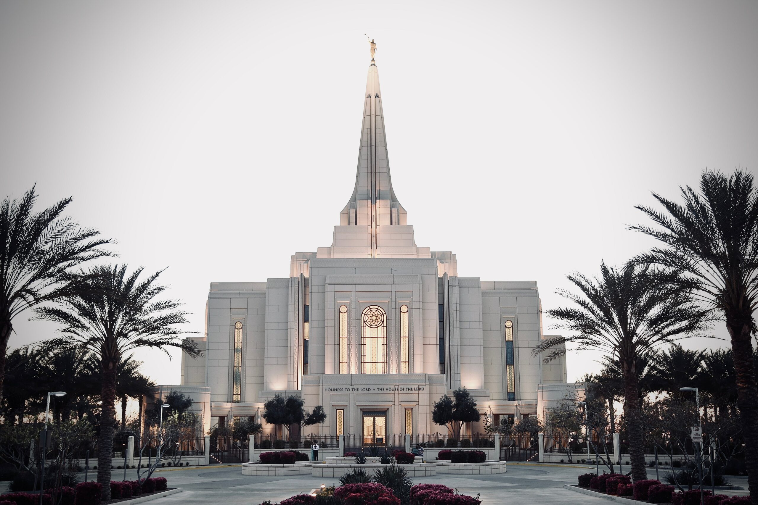 A white stone building with a tall spire and angel statue on top, surrounded by palm trees and landscaped gardens.