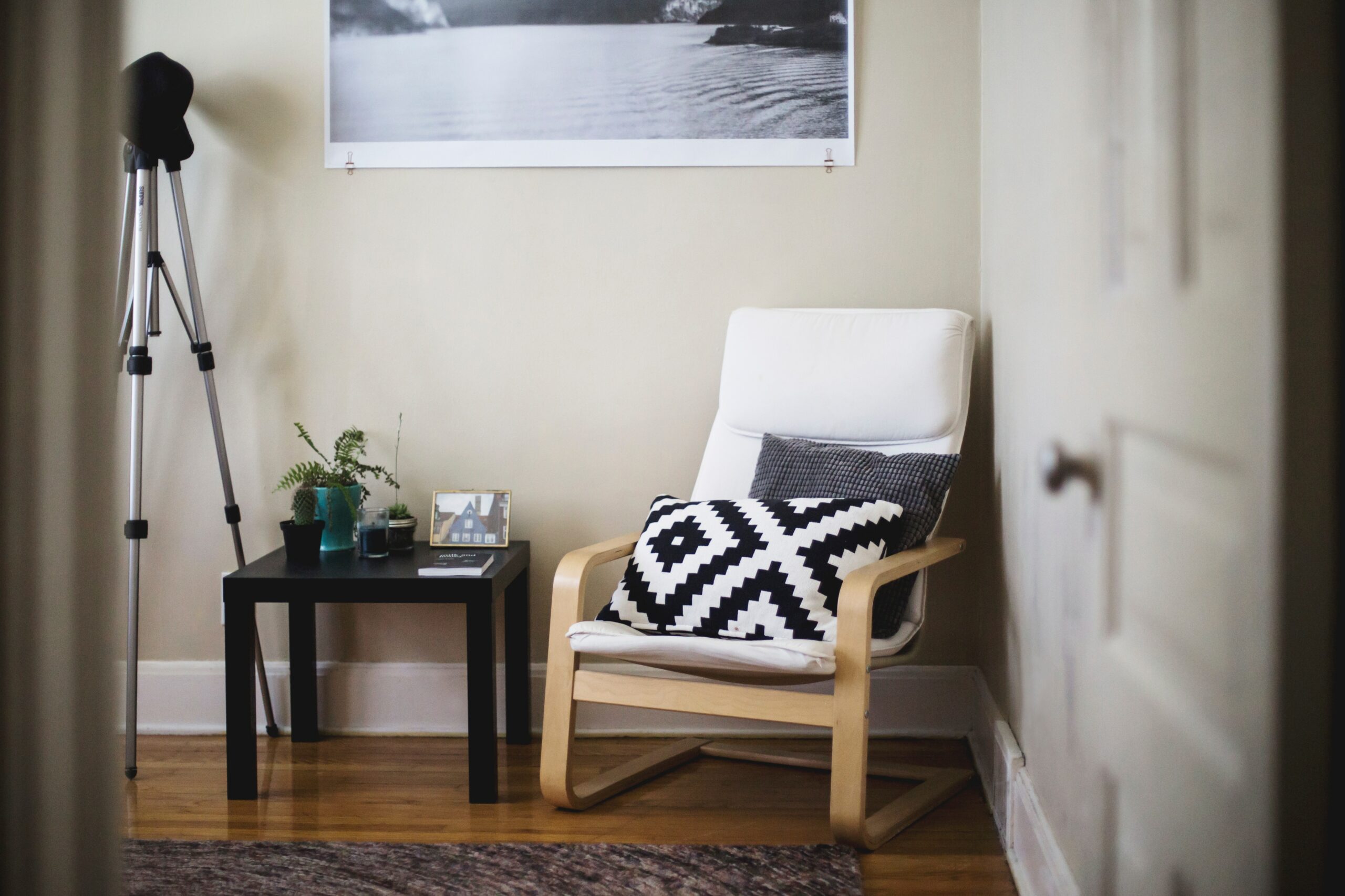 A modern reading nook with a white armchair, two patterned cushions, a black side table with decor, a plant, a tripod, and a black-and-white wall photo.