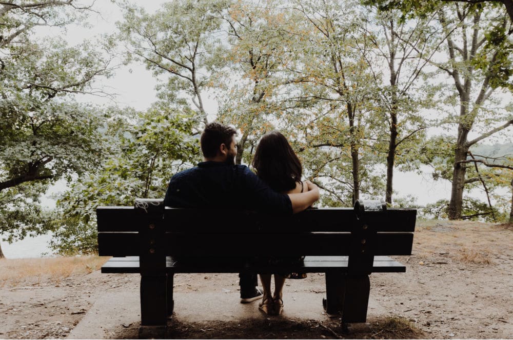 Two people sit closely together on a park bench, viewed from behind, surrounded by trees and overlooking a natural outdoor setting.