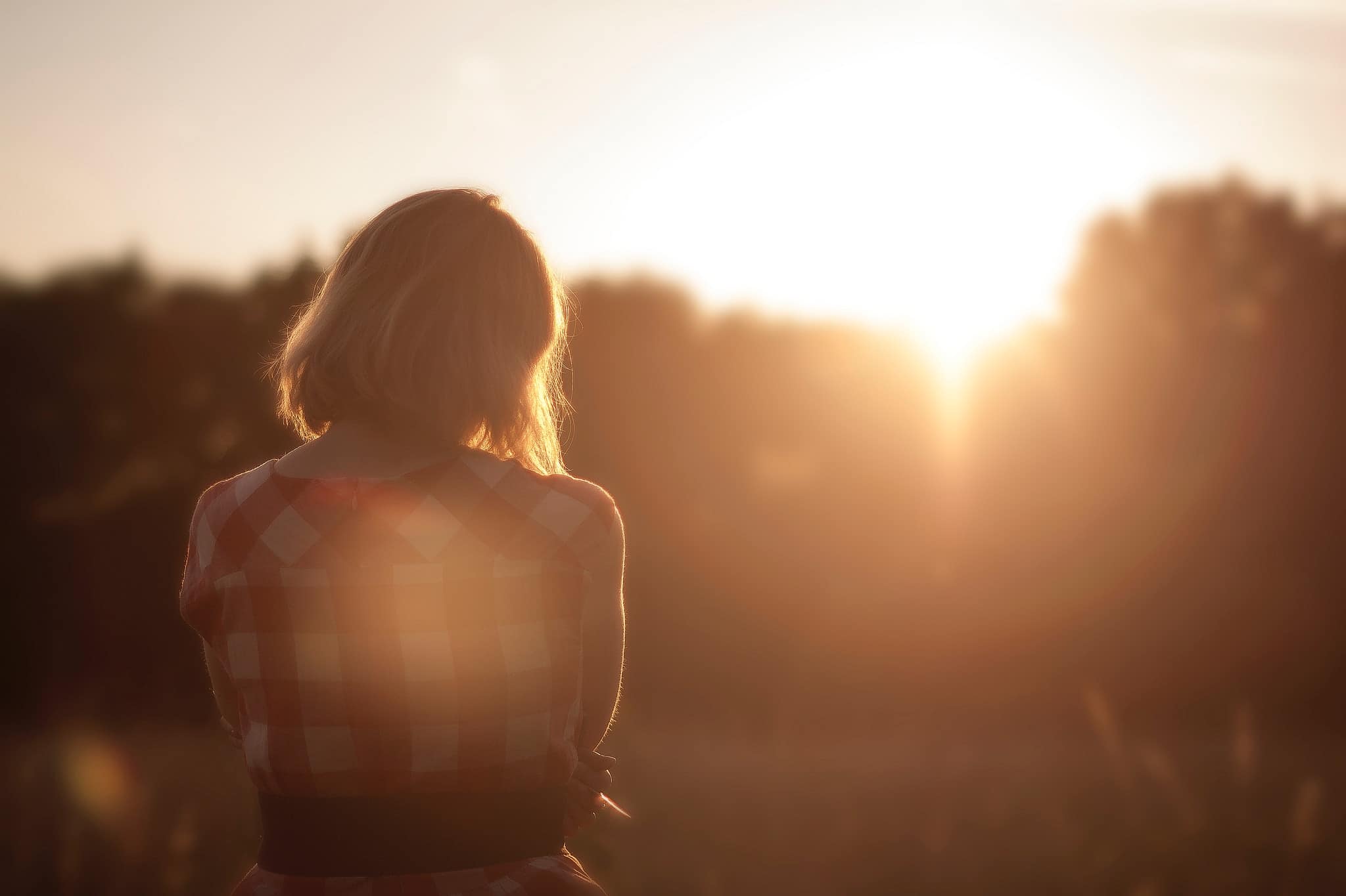 Person with short hair in a plaid shirt stands outdoors at sunset, facing away from the camera toward the bright sun and blurred trees.