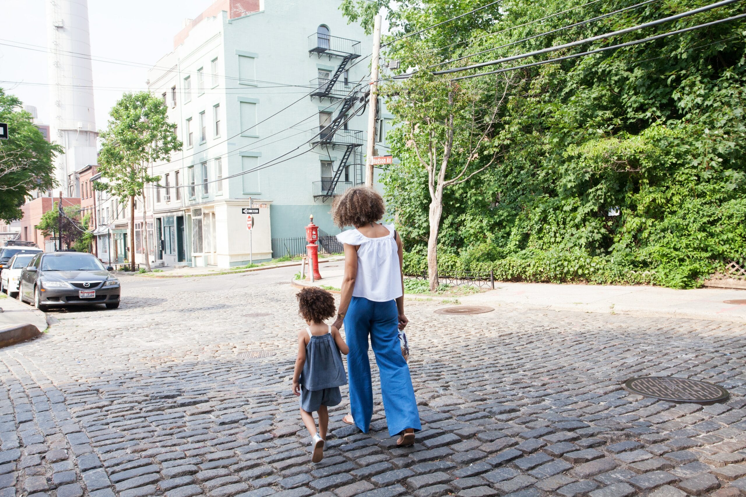 A woman and a young girl holding hands walk across a cobblestone street in a city neighborhood on a sunny day.