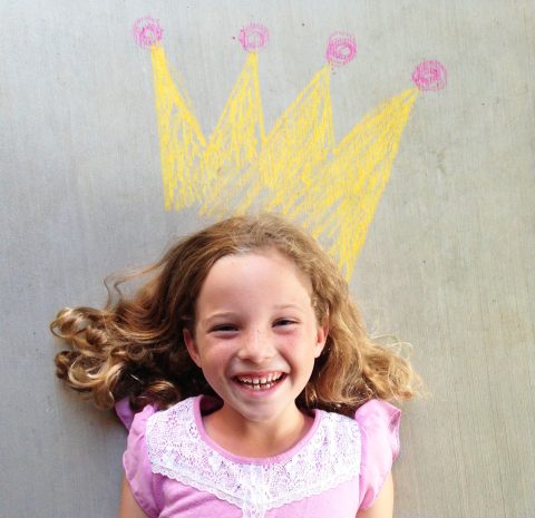 Smiling girl lying on the ground with a yellow crown drawn in chalk above her head, appearing as if she is wearing it.